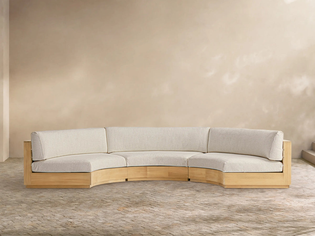 Bal Harbour Outdoor Curved Three Piece Sectional sofa with light wood frame & light beige cushions on a patio with a tan wall.