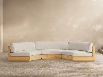 Bal Harbour Outdoor Curved Three Piece Sectional sofa with light wood frame & light beige cushions on a patio with a tan wall.