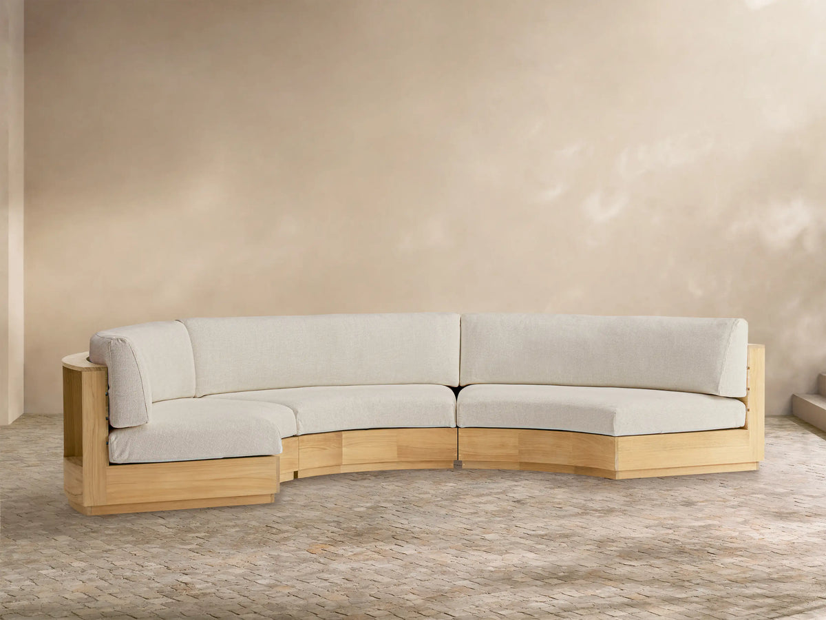 Bal Harbour Outdoor Curved Three Piece Sectional sofa with light wood frame & light beige cushions on a patio with a tan wall.