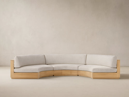 Bal Harbour Outdoor Curved Three Piece Sectional sofa with light wood frame & light beige cushions on a patio with a tan wall.