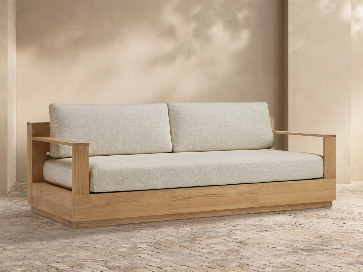 Bal Harbour Outdoor Teak Sofa. Natural teak frame with wide armrests and thick light beige cushions on textured stone paving.