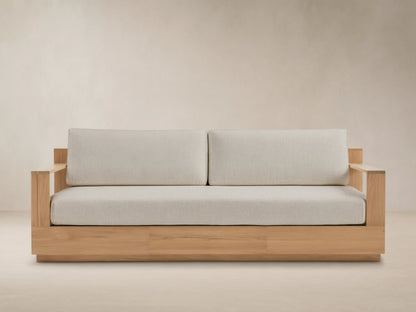 Bal Harbour Outdoor Teak Sofa. Natural teak frame with wide armrests and thick light beige cushions on textured stone paving.