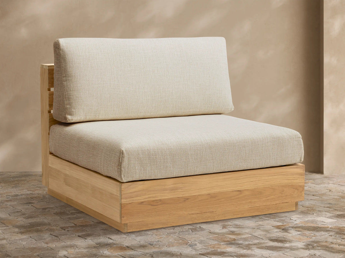 Bal Harbour Outdoor Teak Armless Chair with light natural wood base, light beige seat & back cushions, on a stone patio.