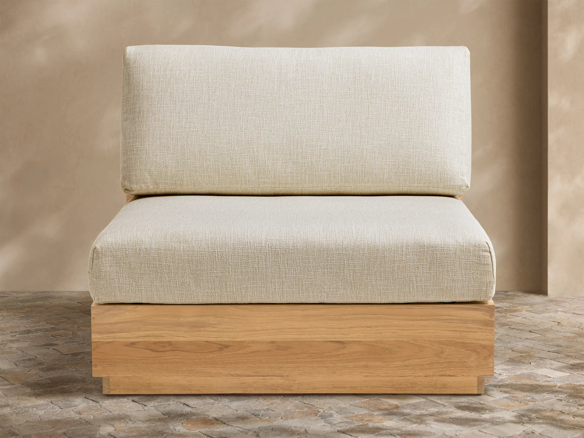 Bal Harbour Outdoor Teak Armless Chair with light natural wood base, light beige seat & back cushions, on a stone patio.