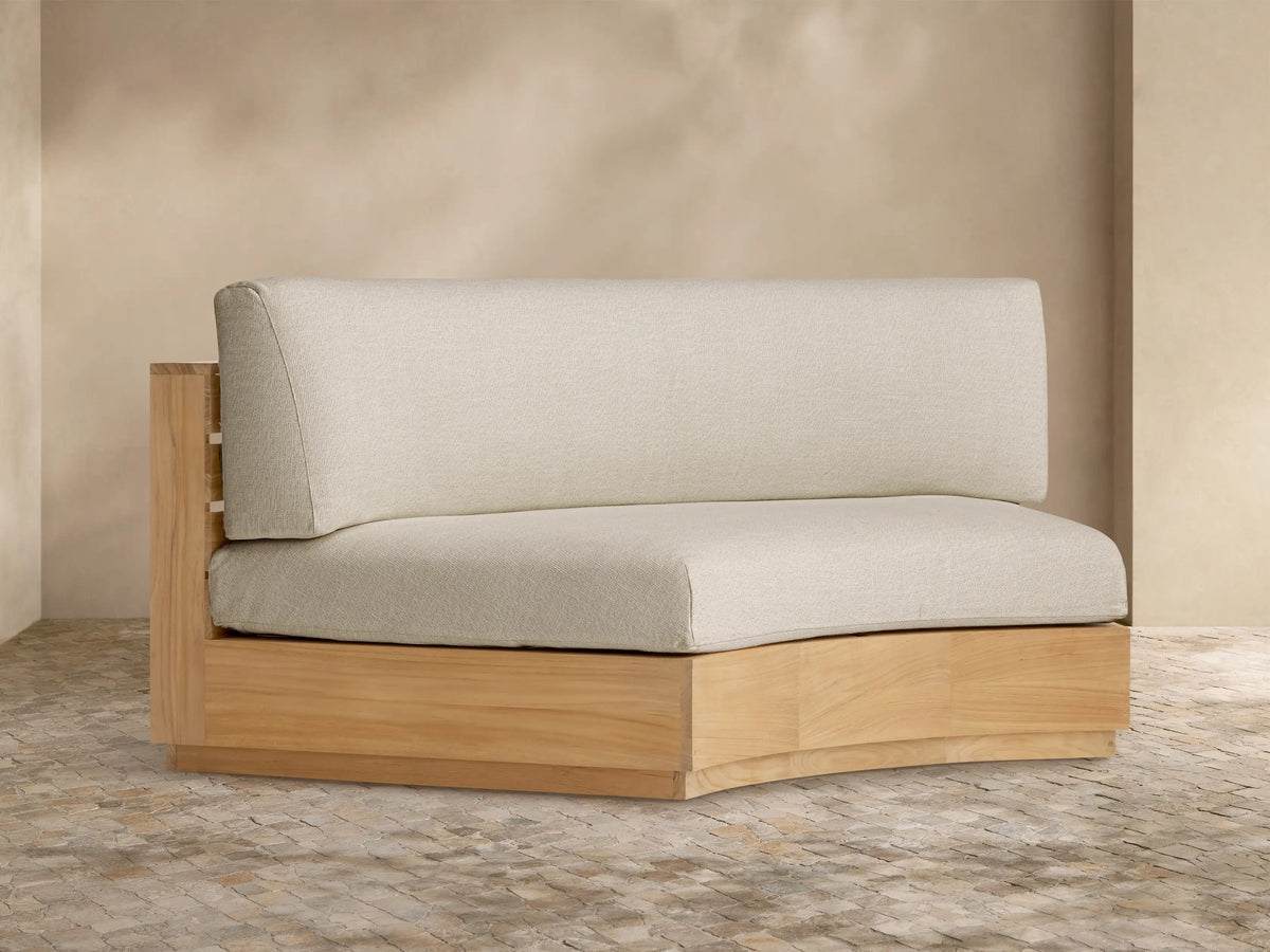Bal Harbour Outdoor Curved Sofa with a natural teak wood base and light beige cushions, shown against a light wall on a stone floor.
