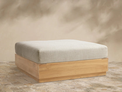 Bal Harbour Outdoor Teak Ottoman with a light grey textured cushion and a natural golden teak wood base on a stone patio.