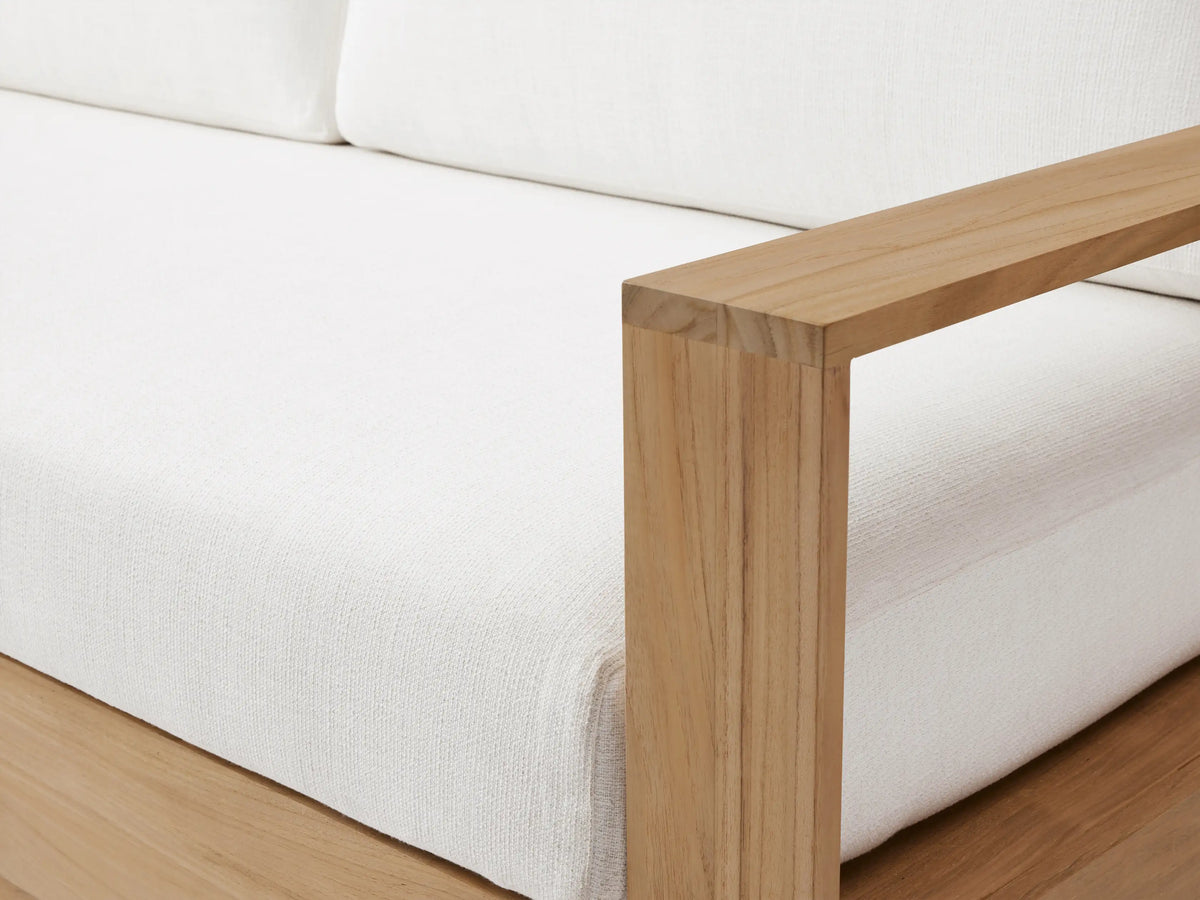 Bal Harbour Outdoor Teak Sofa, natural teak wood frame, white cushions. Viewed angled left in an outdoor space with light wall and paved floor.