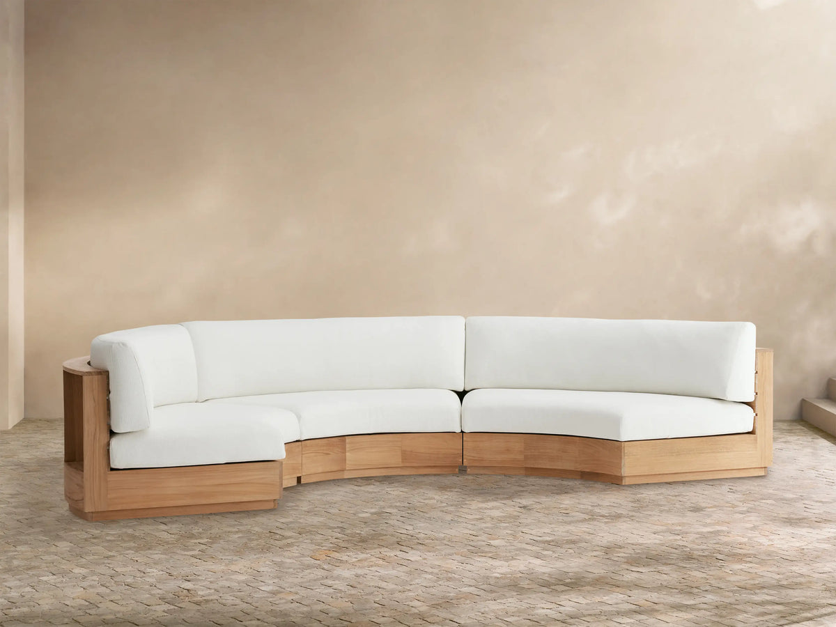 Bal Harbour Outdoor Curved Three Piece Sectional with light wood frame & white cushions on a stone patio against a tan wall.