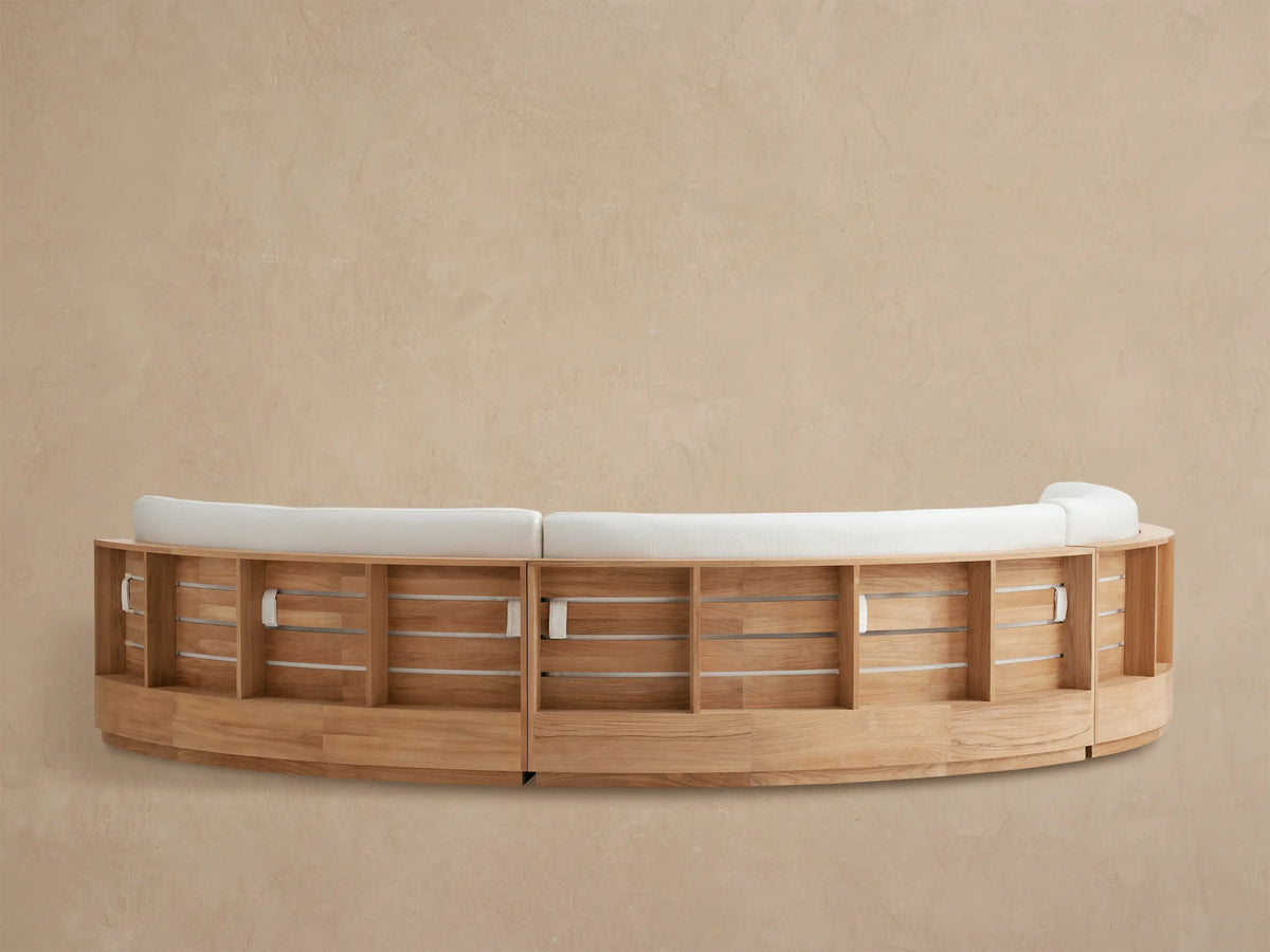 Close-up of Bal Harbour Outdoor Curved Sectional back. Light wood frame with curved slats supports a thick, light beige cushion.
