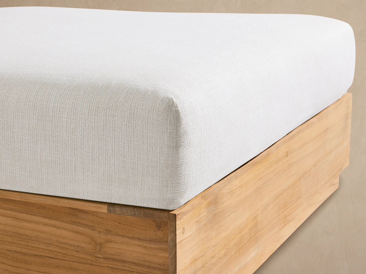 Bal Harbour Outdoor Teak Ottoman with a light natural teak wood base and a thick, soft white cushion, on a stone floor.