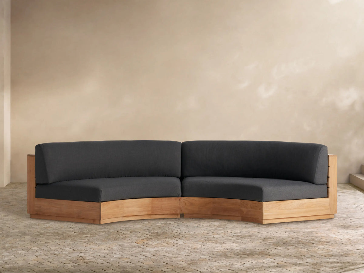 Bal Harbour Outdoor Curved Two-Piece Sectional with natural teak base and charcoal cushions on a paved outdoor patio.