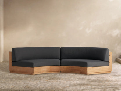 Bal Harbour Outdoor Curved Two-Piece Sectional with natural teak base and charcoal cushions on a paved outdoor patio.
