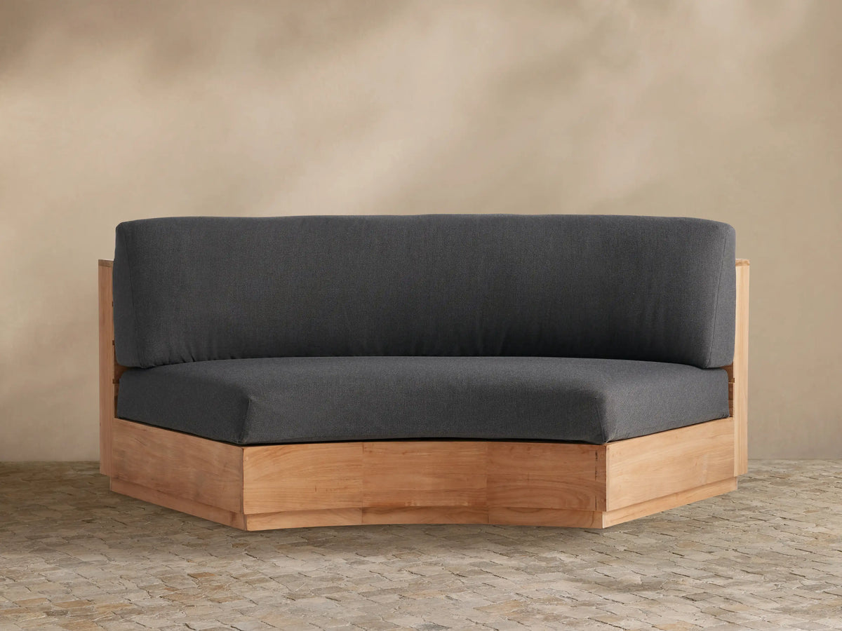 Bal Harbour Outdoor Curved Sofa in natural teak wood, featuring dark charcoal gray cushions, sitting on a stone patio.