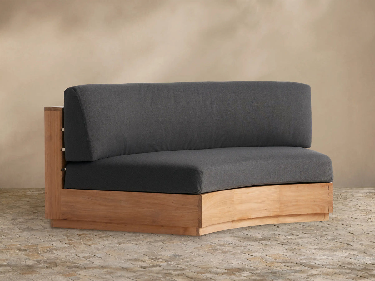 Bal Harbour Outdoor Curved Sofa in natural teak wood, featuring dark charcoal gray cushions, sitting on a stone patio.