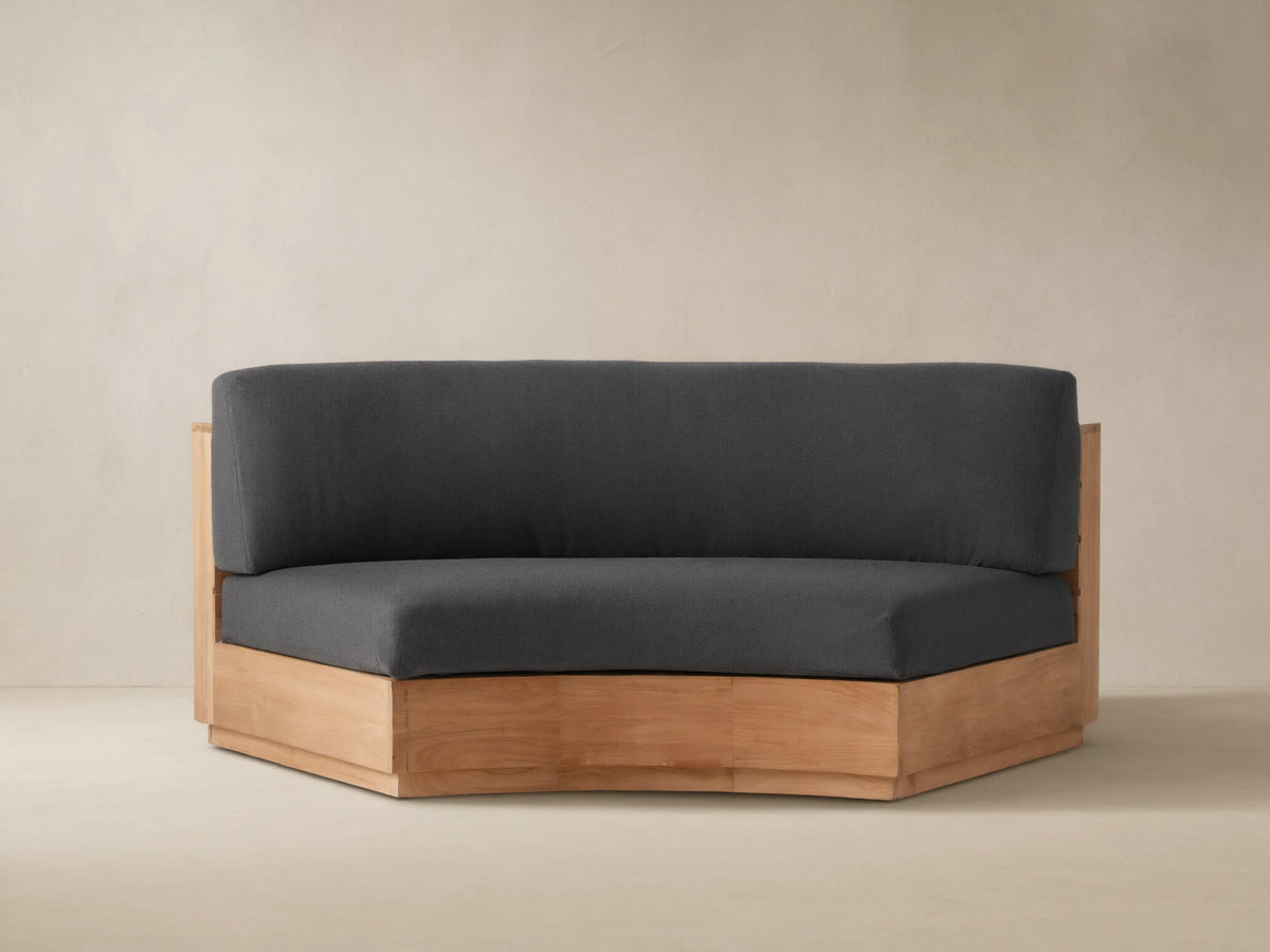 Bal Harbour Outdoor Curved Sofa in natural teak wood, featuring dark charcoal gray cushions, sitting on a stone patio.