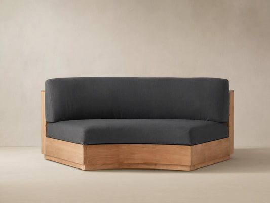 Bal Harbour Outdoor Curved Sofa in natural teak wood, featuring dark charcoal gray cushions, sitting on a stone patio.
