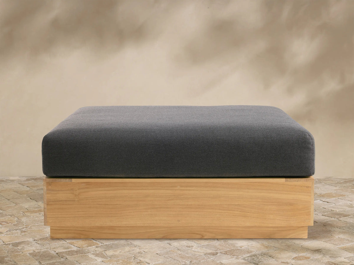 Front view of Bal Harbour Outdoor Teak Ottoman: dark grey cushion on rectangular natural teak wood base, on stone pavers.