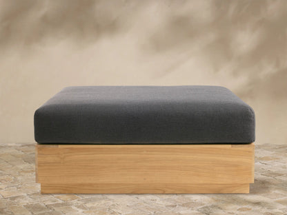 Front view of Bal Harbour Outdoor Teak Ottoman: dark grey cushion on rectangular natural teak wood base, on stone pavers.