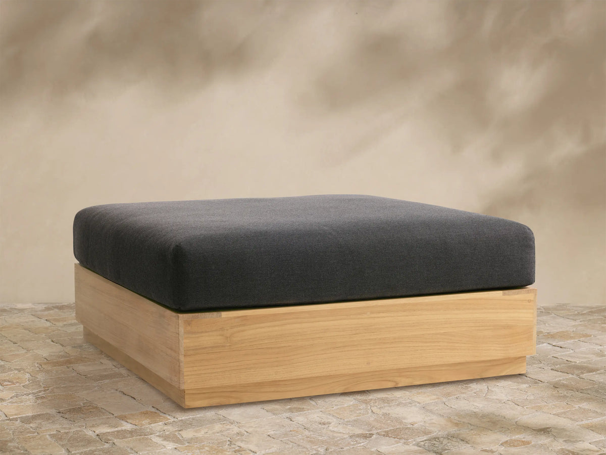 Front view of Bal Harbour Outdoor Teak Ottoman: dark grey cushion on rectangular natural teak wood base, on stone pavers.