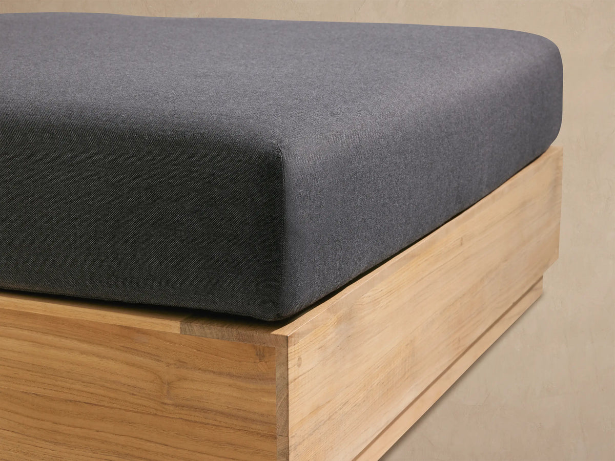 Square Bal Harbour Outdoor Teak Ottoman with dark gray cushion on a light natural teak wood base, set on a stone patio.