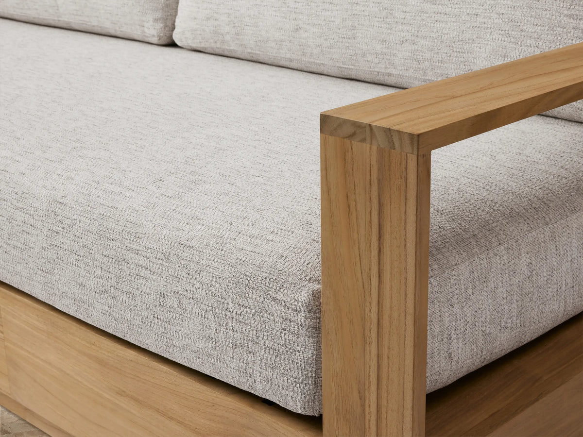 Bal Harbour Outdoor Teak Sofa: modern design, natural golden teak wood frame, light grey fabric cushions. Angled view on a light patio.