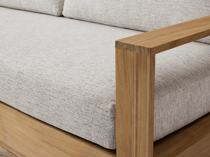 Bal Harbour Outdoor Teak Sofa: modern design, natural golden teak wood frame, light grey fabric cushions. Angled view on a light patio.