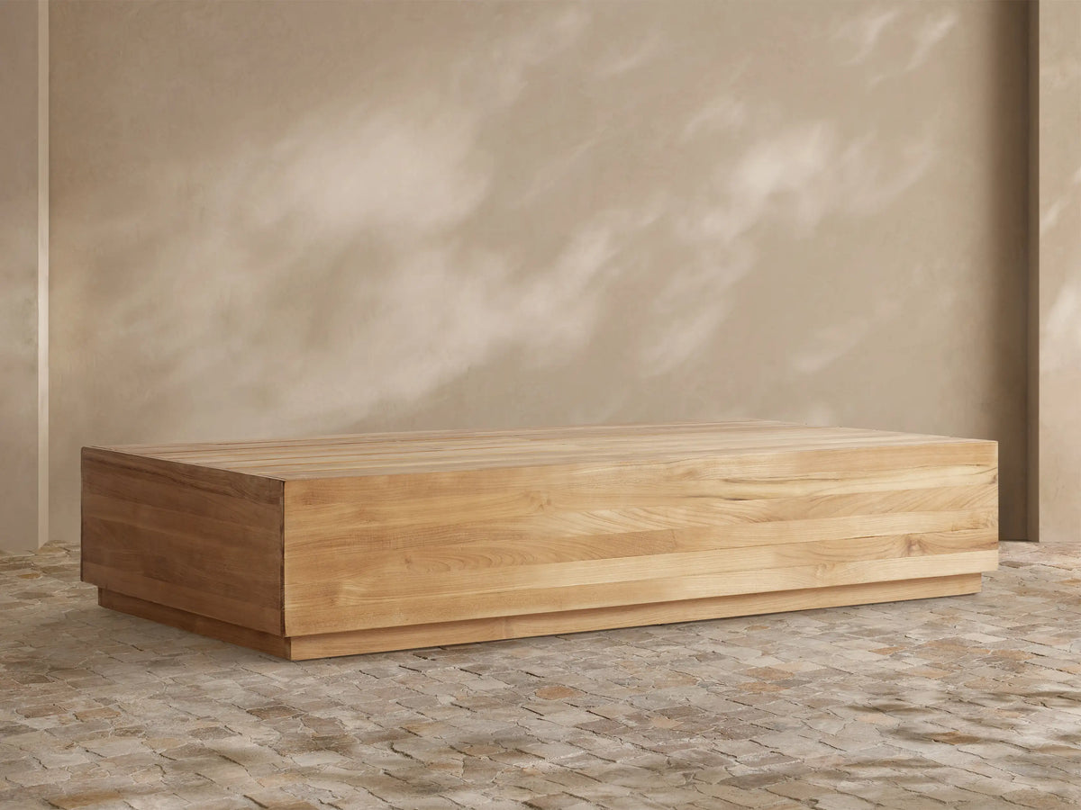 Bal Harbour Outdoor Teak Coffee Table: Rectangular light golden-brown teak table with slatted top on a stone floor, natural light.