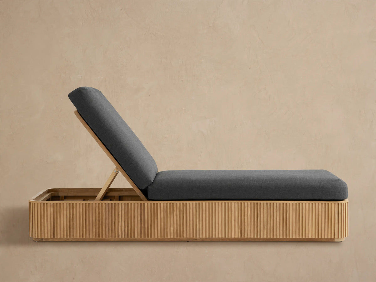 Beaumont Outdoor Teak Chaise with dark gray cushions, inclined back, and slatted light brown teak base, on a paved patio.