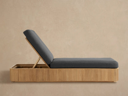 Beaumont Outdoor Teak Chaise with dark gray cushions, inclined back, and slatted light brown teak base, on a paved patio.