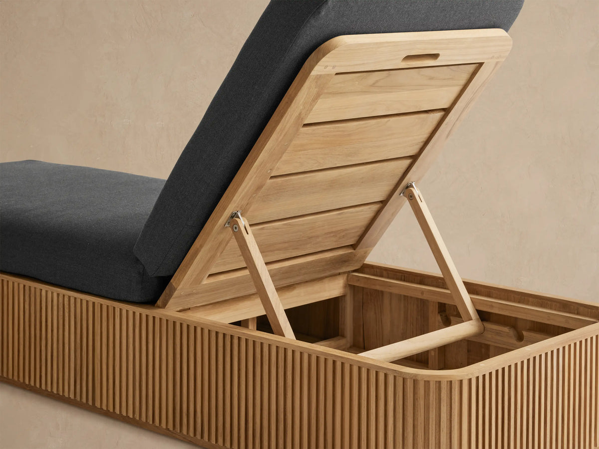 Beaumont Outdoor Teak Chaise, side view. Features a slatted teak wood base and a thick dark gray cushion with an inclined backrest.