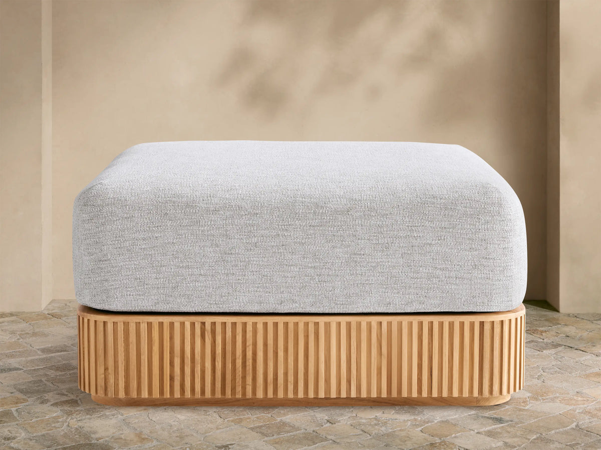 Beaumont Outdoor Teak Ottoman with a light gray cushion on a slatted natural teak wood base, outdoors on stone pavement.