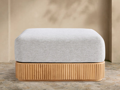 Beaumont Outdoor Teak Ottoman with a light gray cushion on a slatted natural teak wood base, outdoors on stone pavement.