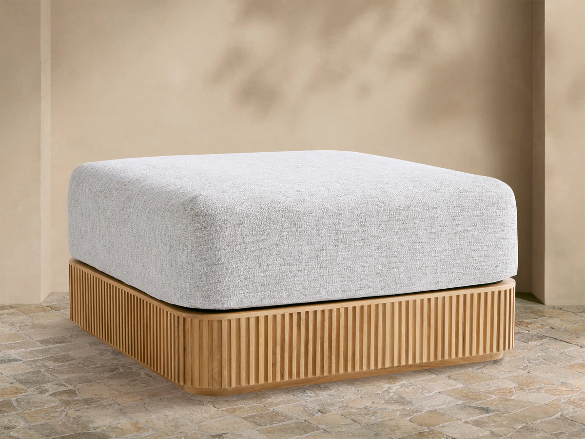 Beaumont Outdoor Teak Ottoman with a light gray cushion on a slatted natural teak wood base, outdoors on stone pavement.
