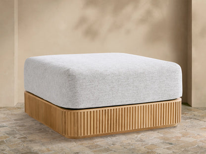 Beaumont Outdoor Teak Ottoman with a light gray cushion on a slatted natural teak wood base, outdoors on stone pavement.