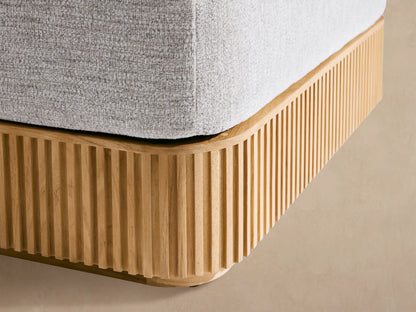 Beaumont Outdoor Teak Ottoman. Light grey cushion on a warm, slatted natural teak base. Seen from front-right on a stone patio.