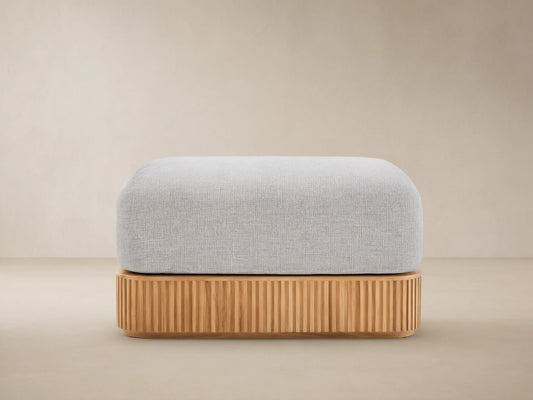 Beaumont Outdoor Teak Ottoman with a light gray cushion on a slatted natural teak wood base, outdoors on stone pavement.