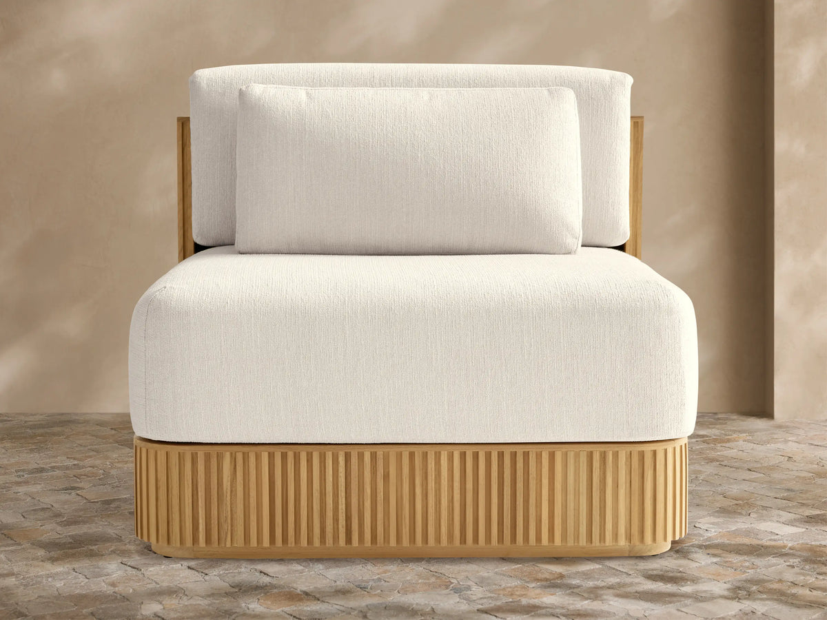 Beaumont Teak Lounge Chair. Front view of modern chair with slatted natural teak base & thick cream cushions on a stone patio.