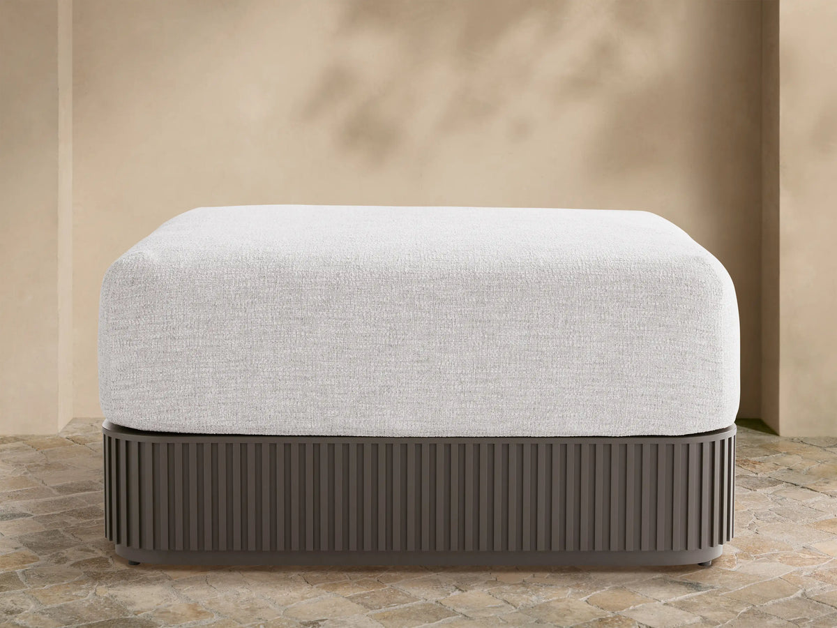 Light grey upholstered Beaumont Outdoor Ottoman with dark grey slatted aluminum base on a stone patio, beige wall background.