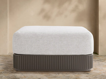 Light grey upholstered Beaumont Outdoor Ottoman with dark grey slatted aluminum base on a stone patio, beige wall background.