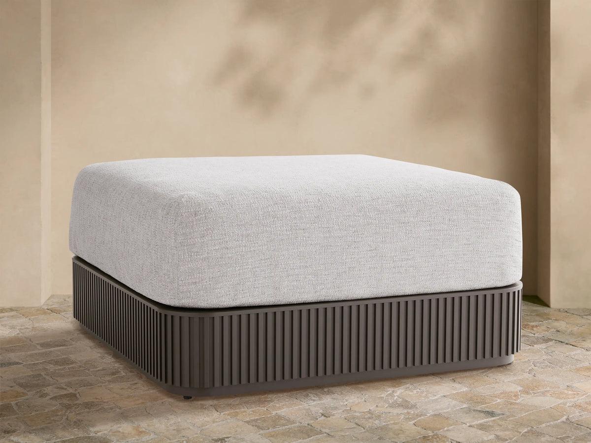 Light grey upholstered Beaumont Outdoor Ottoman with dark grey slatted aluminum base on a stone patio, beige wall background.