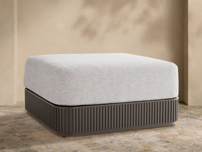 Light grey upholstered Beaumont Outdoor Ottoman with dark grey slatted aluminum base on a stone patio, beige wall background.