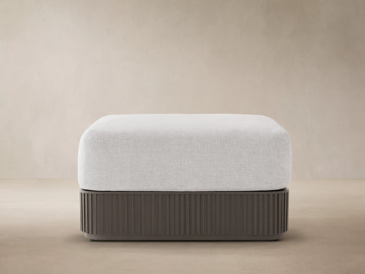 Light grey upholstered Beaumont Outdoor Ottoman with dark grey slatted aluminum base on a stone patio, beige wall background.