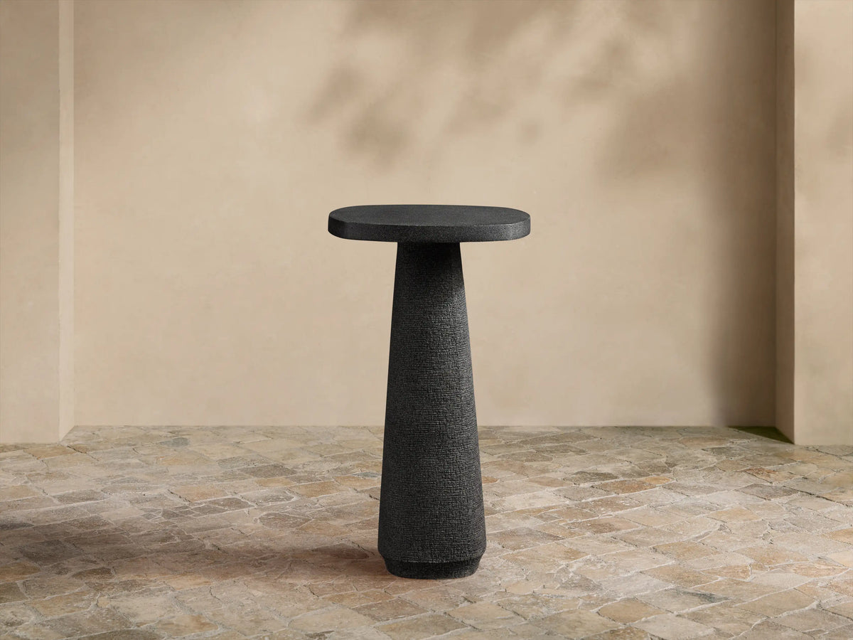 Bellagio Outdoor End Table: Dark gray, textured, with an oval top and tapered column base, on a stone patio against a light wall.