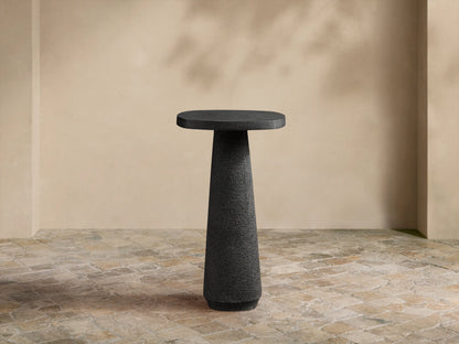 Bellagio Outdoor End Table: Dark gray, textured, with an oval top and tapered column base, on a stone patio against a light wall.