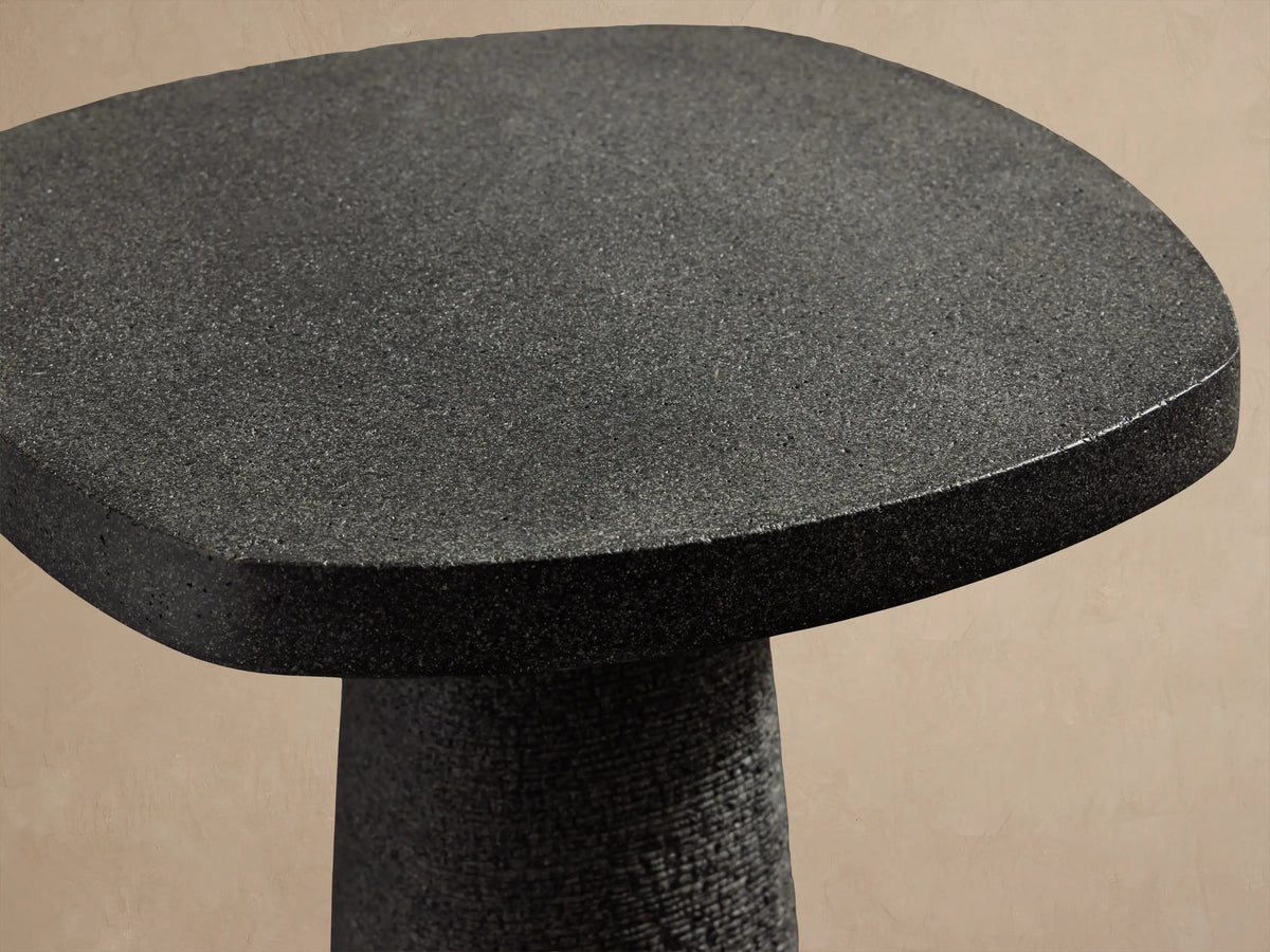 Bellagio Outdoor End Table: Dark gray, textured, with an oval top and tapered column base, on a stone patio against a light wall.