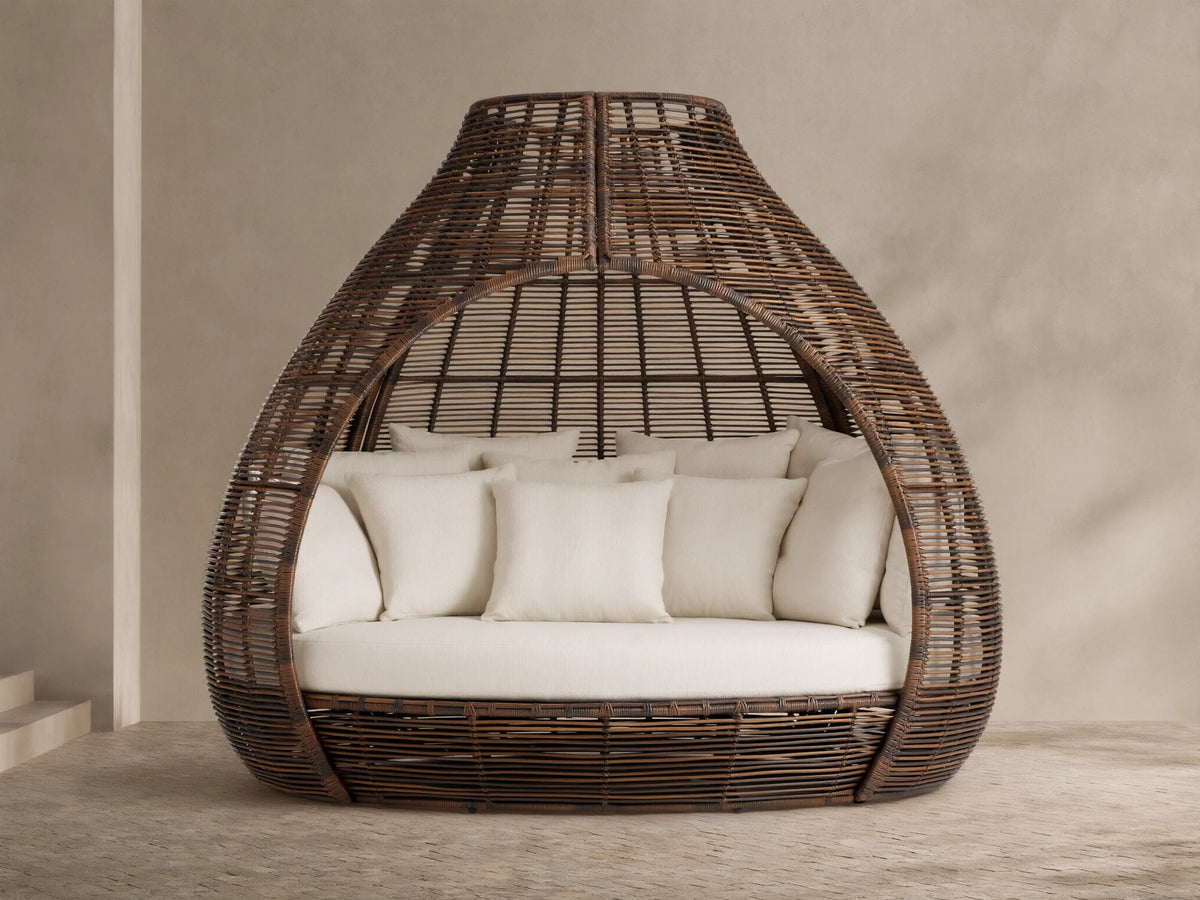 Front view of the dark brown woven Calusa Outdoor Daybed with an open, tapered dome design, filled with cream seat and toss cushions.