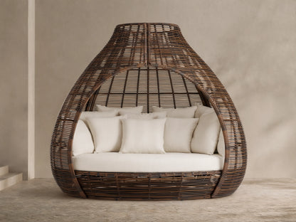 Front view of the dark brown woven Calusa Outdoor Daybed with an open, tapered dome design, filled with cream seat and toss cushions.