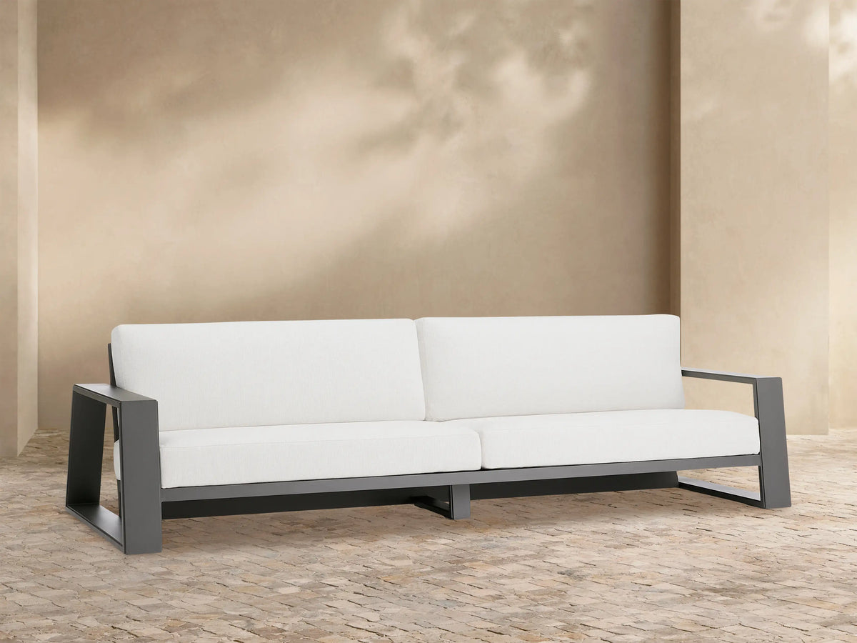 Canyon Outdoor Aluminum Sofa, dark gray frame with white cushions, centered on a stone patio against a light beige wall.