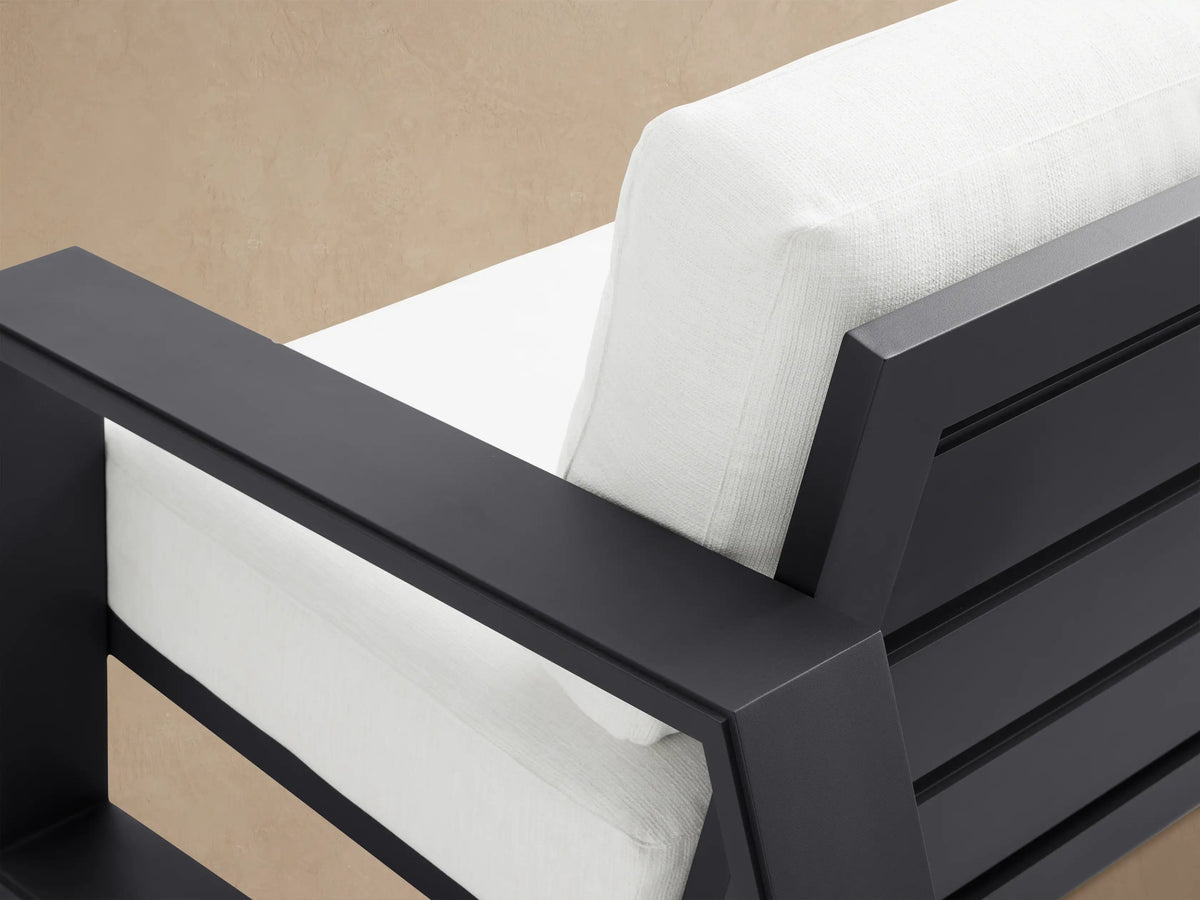 Canyon Outdoor Aluminum Sofa, two-seater, with a sleek dark gray frame & thick white cushions, on a patio with a light wall.