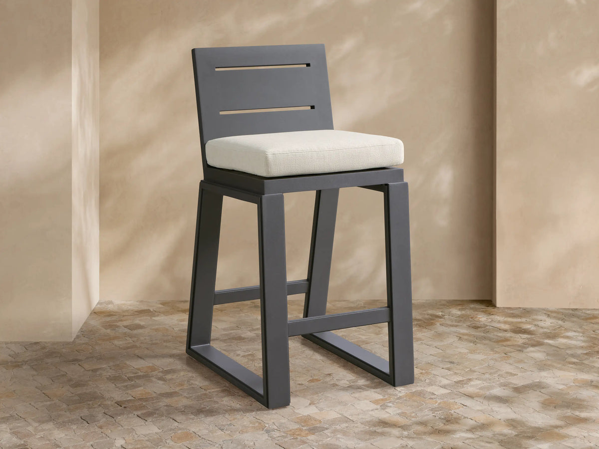 Dark gray Canyon Outdoor Aluminum Barstool with a slatted back and light beige cushion, on a mosaic patio with textured beige walls.